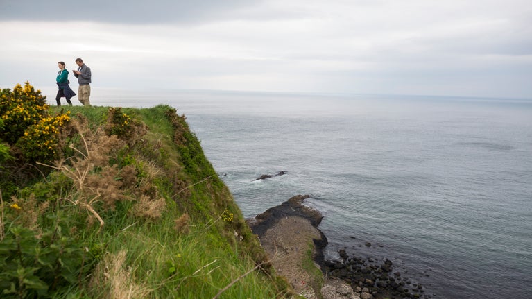 Two people standing on top of a cliff with the sea in the background